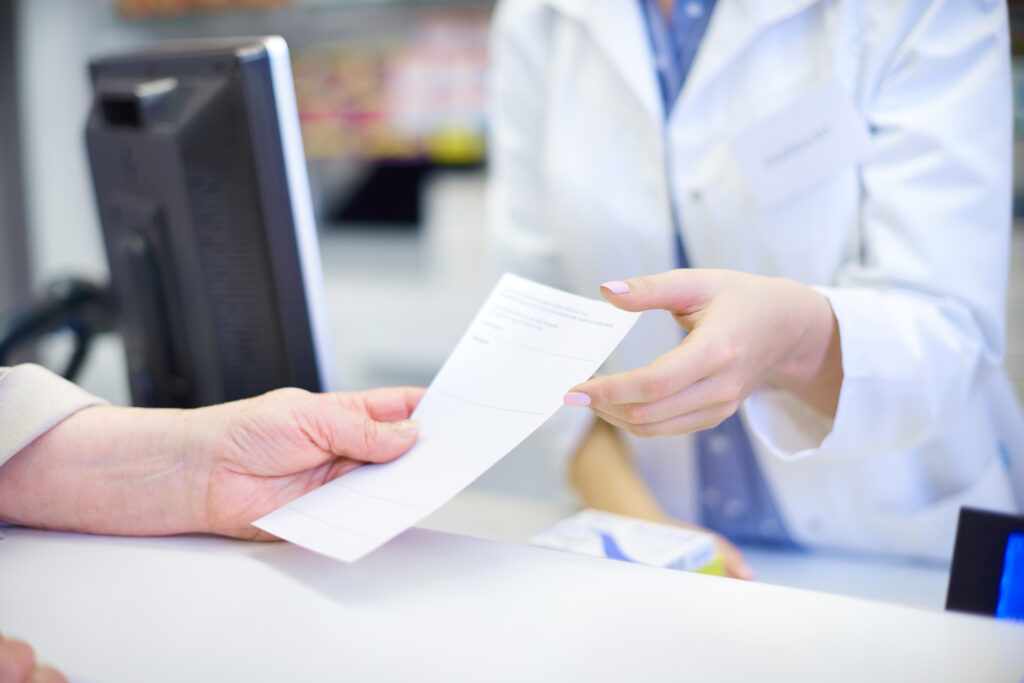 A close-up image of two individuals at a pharmacy counter, where one person is handing over a prescription paper to another whose attire suggests they are a pharmacist. This image is suitable for the refill prescription page as it represents the common action of submitting a prescription for medication refills.