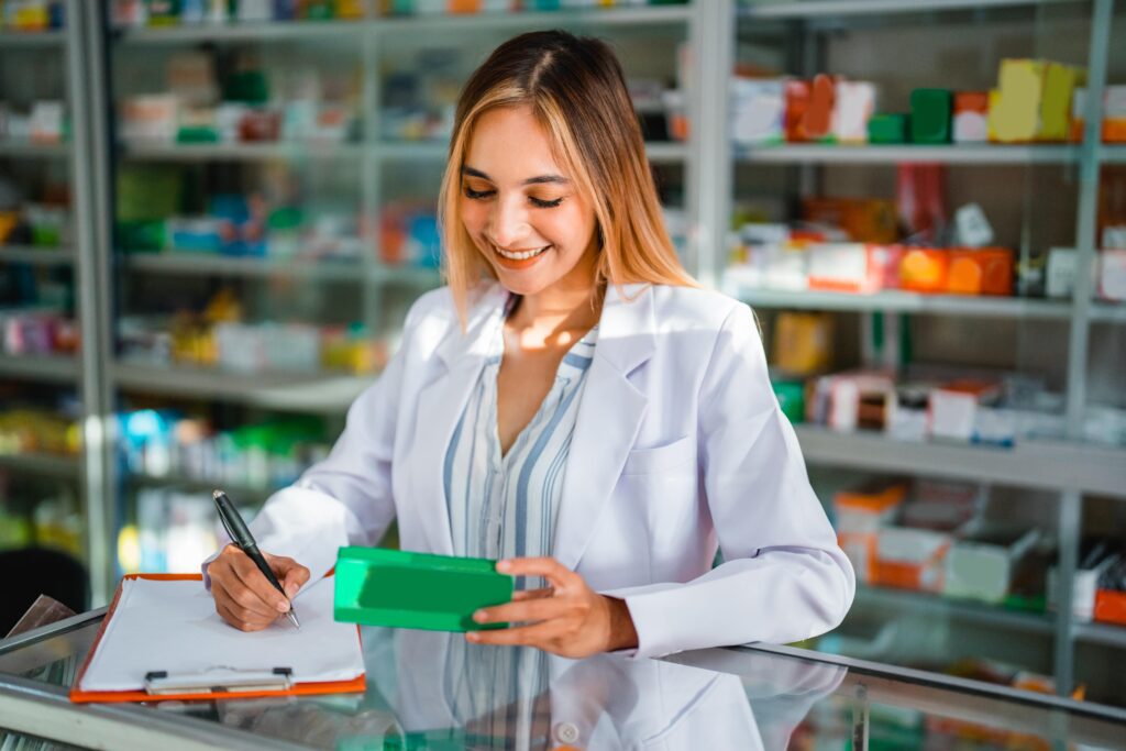 An individual in a white lab coat stands behind a pharmacy counter, holding a green medication box and writing on a clipboard. The background is filled with shelves stocked with various pharmaceutical products. The individual’s face is obscured for privacy