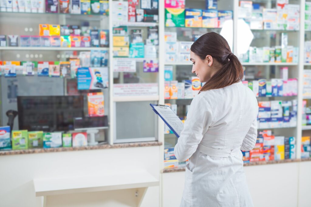 A healthcare professional in a white coat stands in front of a pharmacy counter, holding a clipboard and appearing to take inventory or review patient information. The shelves behind are stocked with various health care products, indicating a well-supplied medical facility. The individual’s identity is obscured for privacy. This image is relevant for illustrating the range of products available at a home health care center and the professionals who manage them.