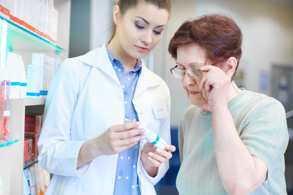 Two individuals in a pharmacy setting, one wearing a white lab coat and holding a syringe, and the other in a green sweater, engaged in a consultation. The background shows pharmacy shelves stocked with various items. Both faces are obscured for privacy