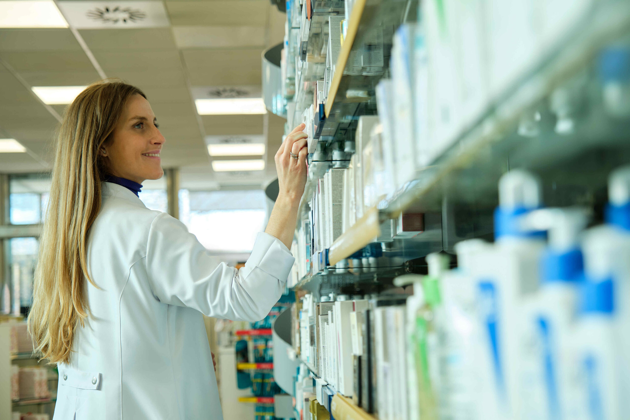 A person in a white lab coat is standing in front of a pharmacy shelf, reaching out to a product. The individual’s face is obscured for privacy. The shelves are stocked with various boxes and medical supplies, indicating the setting is a pharmacy