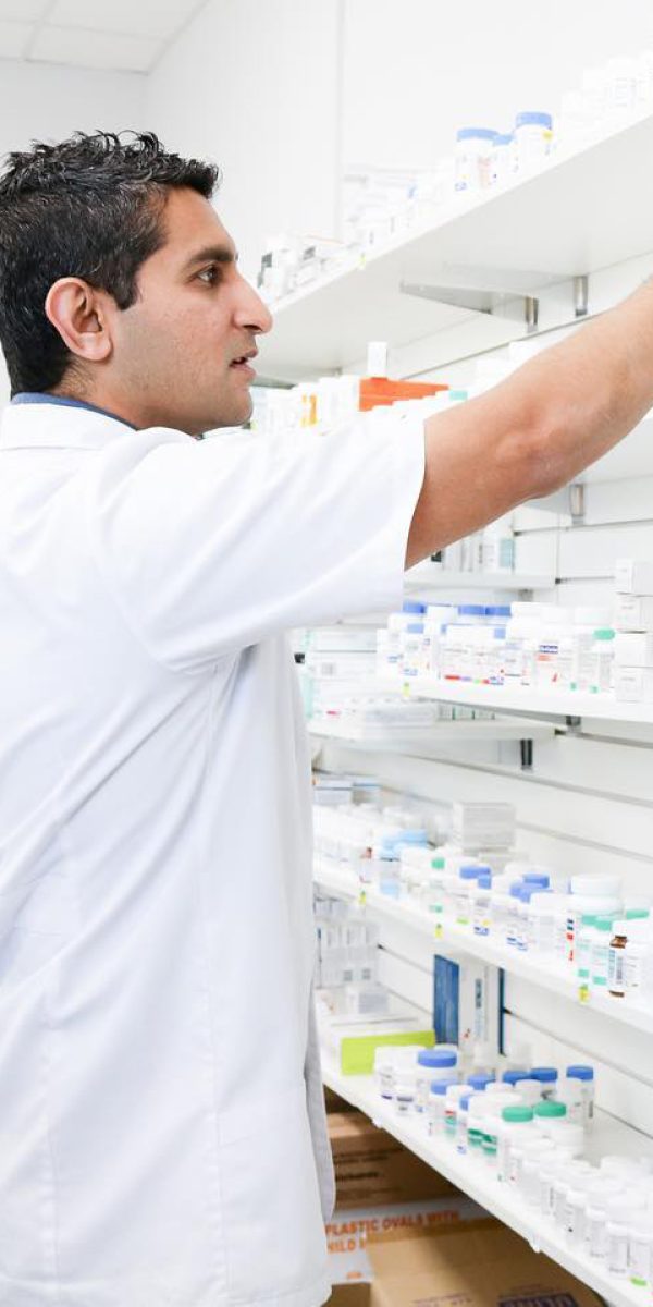 A person in a white coat, presumably a pharmacist, is reaching for a medication on a shelf in Universal Health Pharmacy in Calgary. The pharmacy shelves are stocked with various medications and health products, indicating a well-organized and equipped facility.